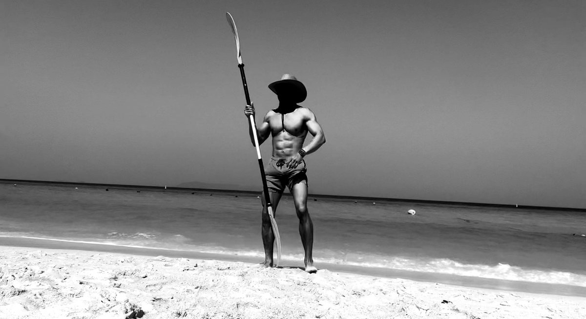 Masked figure standing on a beach holding a paddle vertically.